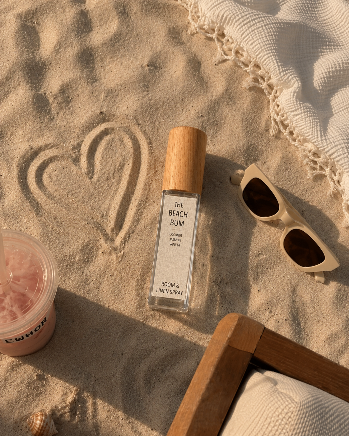 A bottle of 'The Beach Bum' room and linen spray stands on a sandy beach next to a heart drawn in the sand, beige sunglasses, a drink, and a white woven blanket.
